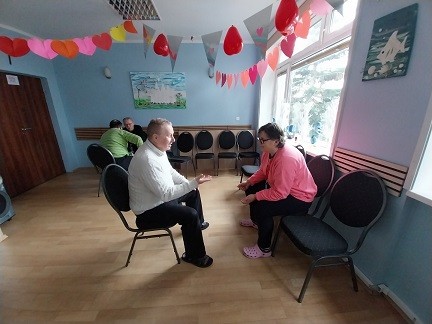 : W udekorowanej sali kilka par uczestników siedzi naprzeciw siebie na krzesłach i prowadzi spokojne rozmowy podczas zajęć.