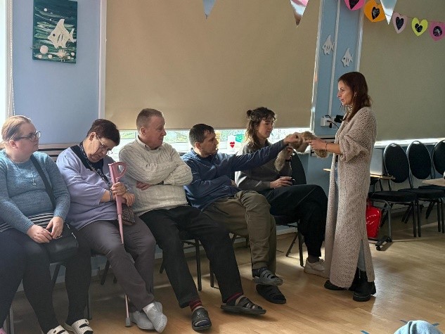 Grupa osób siedzi w sali na krzesłach, a jedna osoba stojąca podaje futerkowy ogon osobie w pierwszym rzędzie.