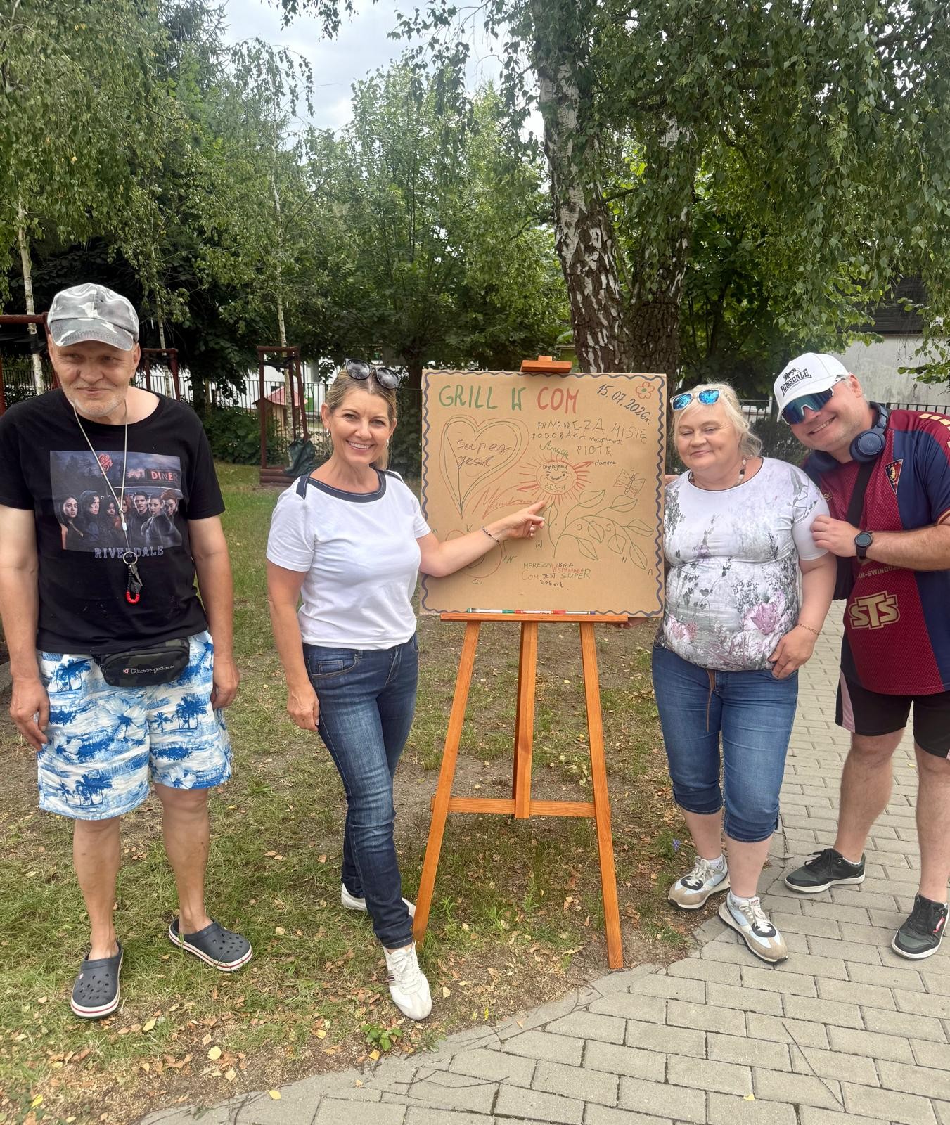 W ogrodzie na sztaludze zawieszony jest plakat z napisem „GRILL W COM”. Po obu bokach sztalugi  stoją cztery  uśmiechnięte osoby .