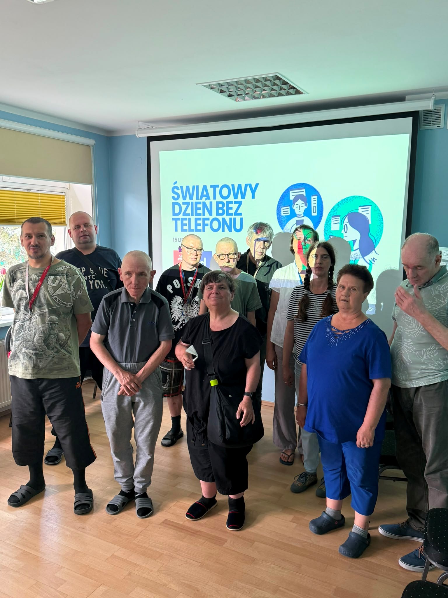 Duża grupa osób stoi w sali przed ekranem, na którym wyświetlany jest napis „ŚWIATOWY DZIEŃ BEZ TELEFONY”.