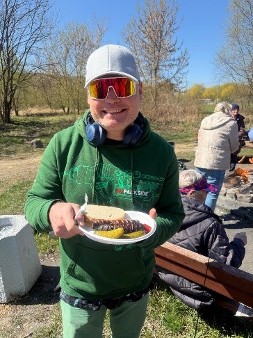 Uśmiechnięty mężczyzna w czapeczce z daszkiem i okularach trzyma w ręku tackę z chlebem i upieczoną kiełbasą. W tle osoby siedzą przy ognisku, widać zieleń i błękit nieba.