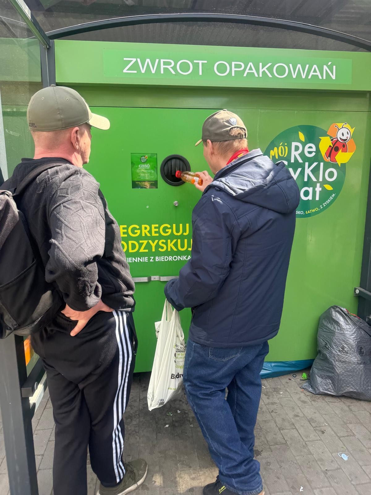 Przed zielonym automatem do recyklingu z napisem „ZWROT OPAKOWAŃ”, stoi dwóch mężczyzn. Ten po prawej wkłada butelkę do otworu.