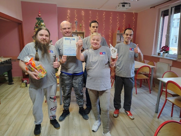 Grupa osób w szarych podkoszulkach prezentuje dyplom, statuetkę i nagrodę w sali ozdobionej świątecznymi dekoracjami.