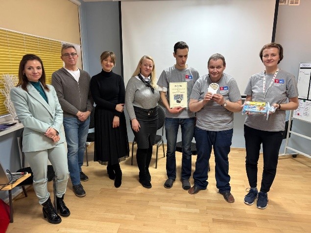 Grupa osób stoi w sali przed ekranem projekcyjnym; część trzyma dyplomy i nagrody, pozując do wspólnego zdjęcia po wydarzeniu.