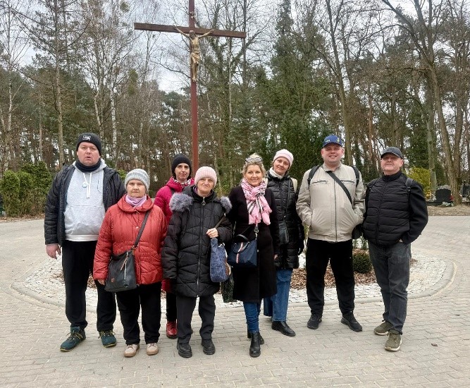 Grupa dziewięciu osób stoi na cmentarzu przy dużym krzyżu. Wszyscy ubrani są w ciepłe kurtki i czapki. W tle widać drzewa i alejki cmentarne. Osoby pozują razem do zdjęcia.