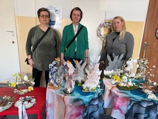 Trzy osoby stoją za stołami przykrytymi kolorowymi tkaninami. Na stołach ułożone są wielkanocne rękodzieła: zajączki, stroiki, wieńce i ozdoby w pastelowych kolorach.