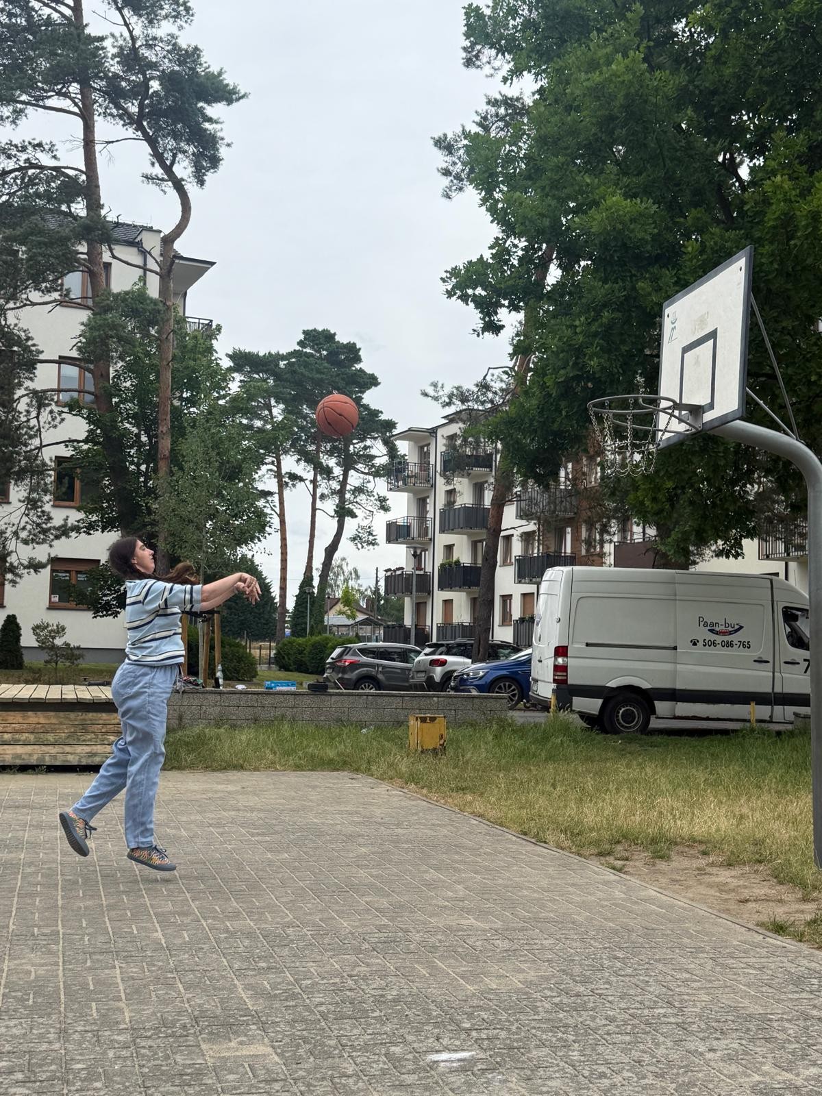 Na boisku kobieta rzuca piłką do kosza. W tle budynki i drzewa.