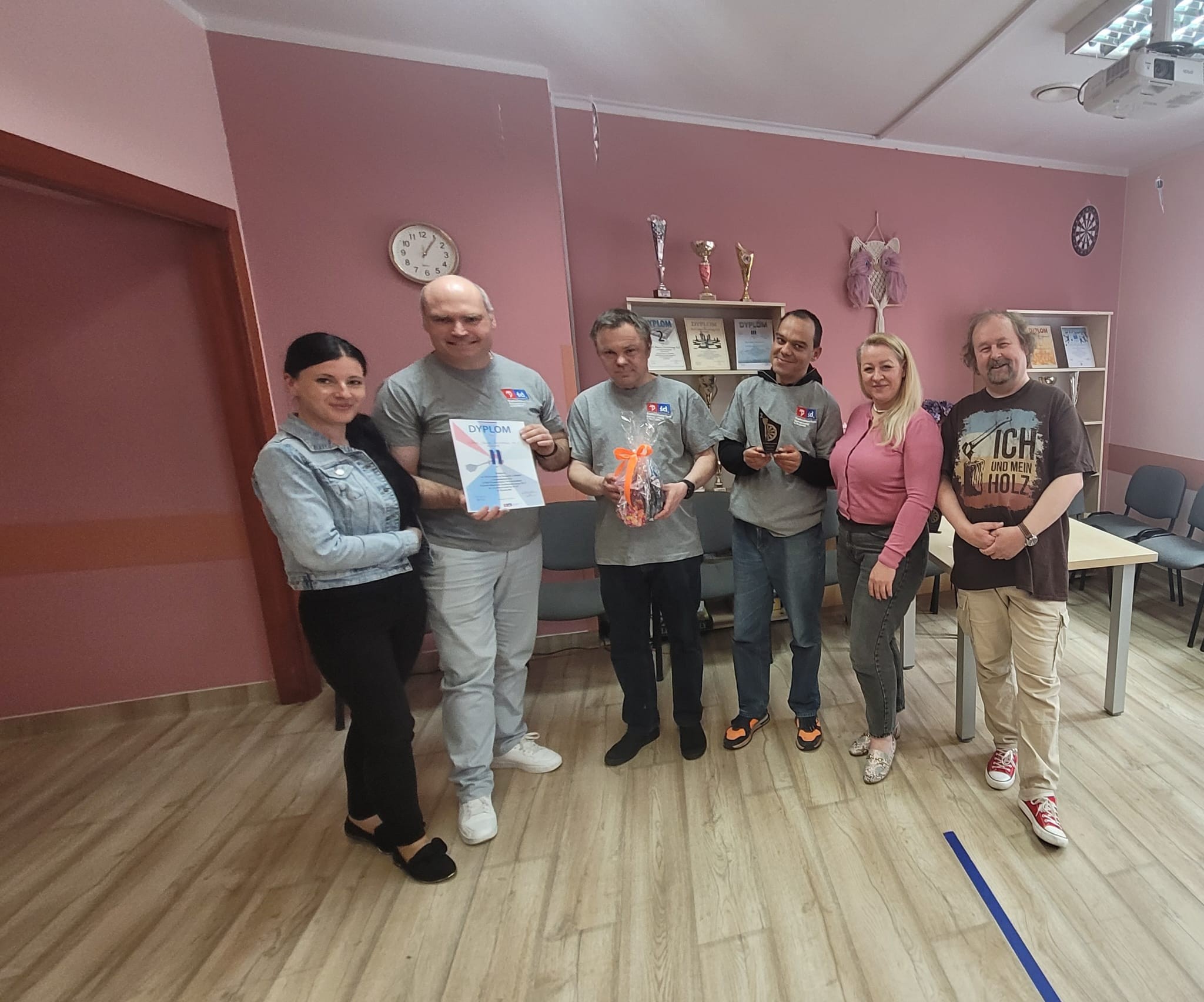 W różowej Sali stoi grupa uśmiechniętych osób pozujących do zdjęcia. Część z nich trzyma dyplom i upominki.