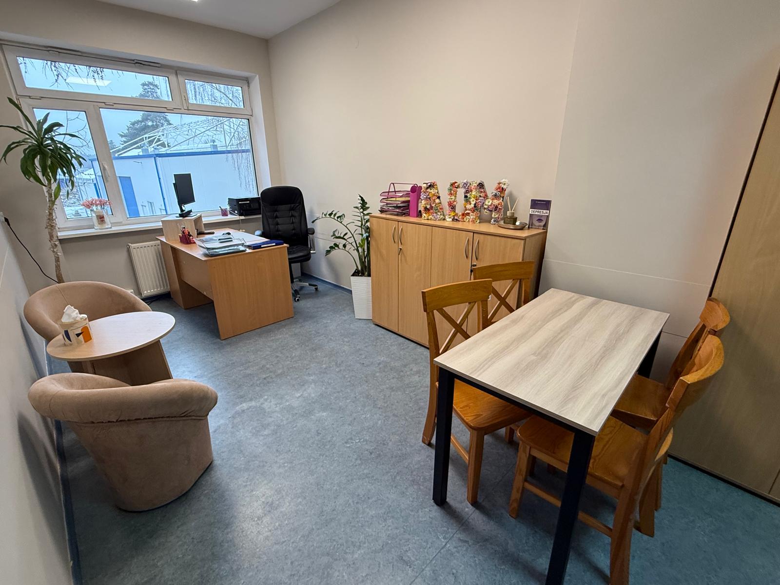 Pokój z oknem,biurko,stół z czterema drewnianymi krzesłami, okrągły stolik oraz fotele.