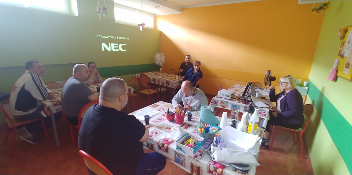 Grupa uczestników ,mężczyzn i kobiet siedzi przy stołach ,kobieta w okularach mówi do nich.