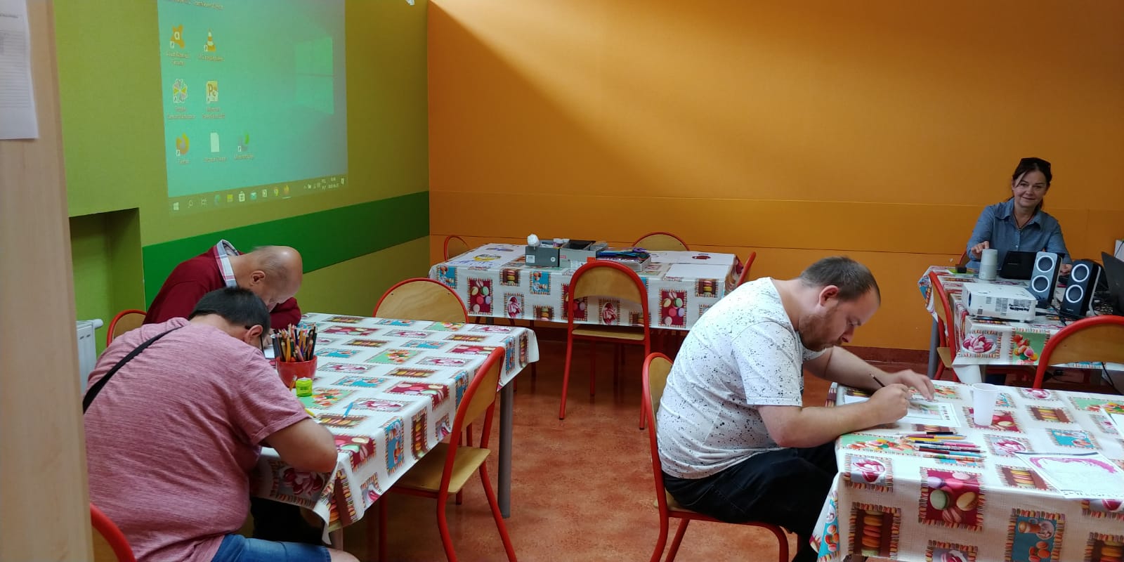 Kobieta siedzi za stołem ,patrzy na trzech siedzących ,pochylonych nad ławkami mężczyzn.