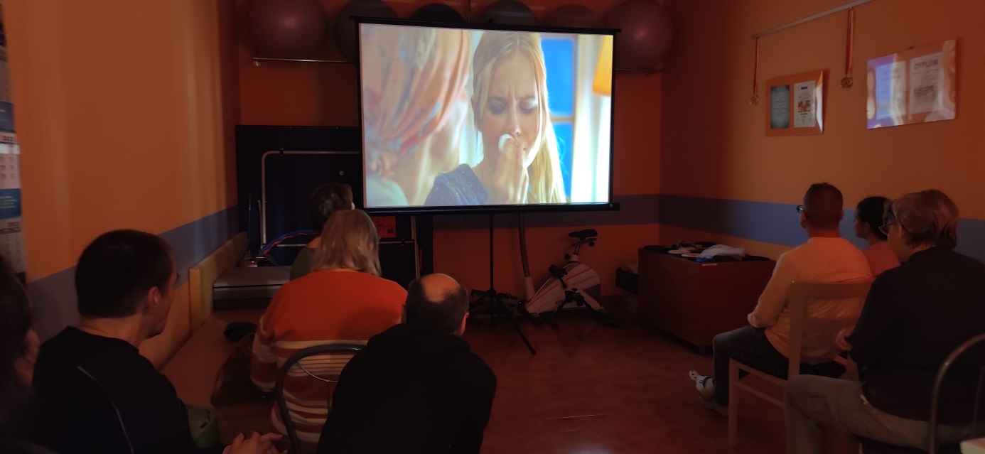 Grupa osób siedzi przed ekranem w sali sportowej.