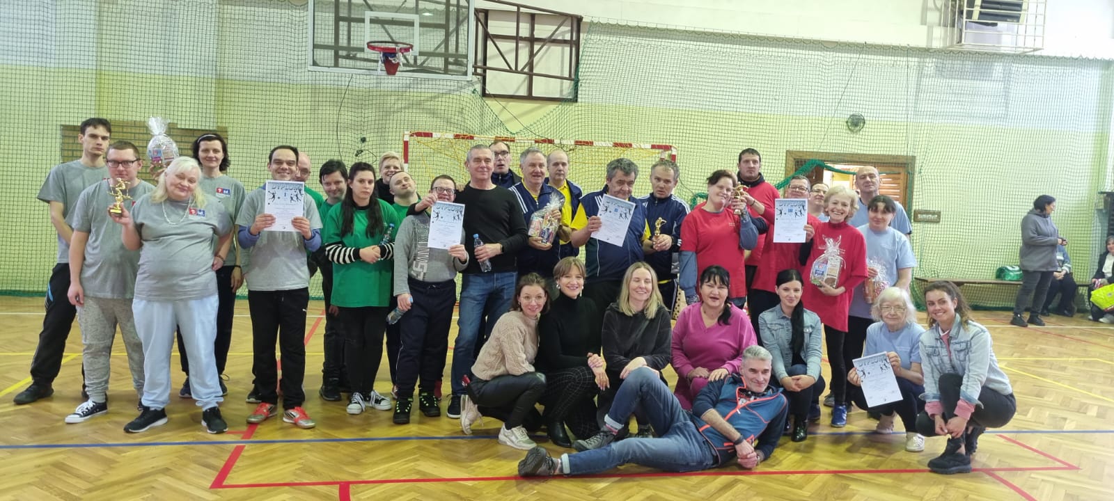 grupa zawodników z dyplomami i pucharami pozuje do zdjecia na hali sportowej.