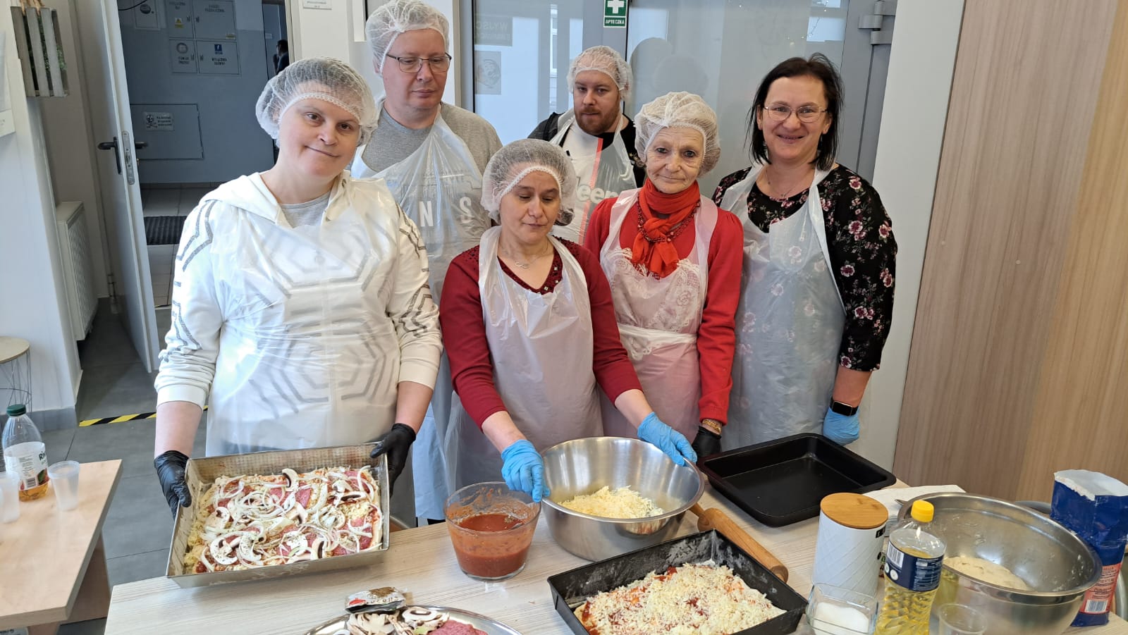Grupa osób w bialych fartuchach stoi przed pizzą na blasze.