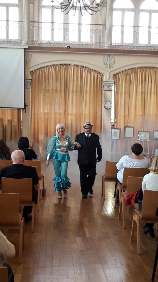 Mężczyzna prowadzi w tańcu kobietę za rękę pomiędzy rzędami krzeseł na dużej hali.