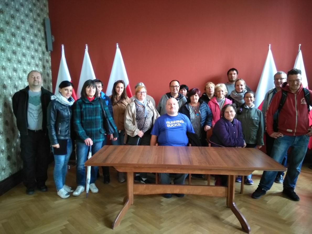 Grupa kobiet i mężczyzn sto za biurkiem,jeden mężczyzna siedz,obok flaga biało –czerwona.