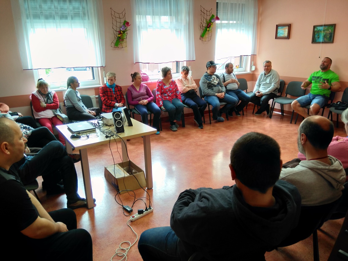 Mężczyzna siedzi pod ścianą  po bokach jego siedzą ludzie.
