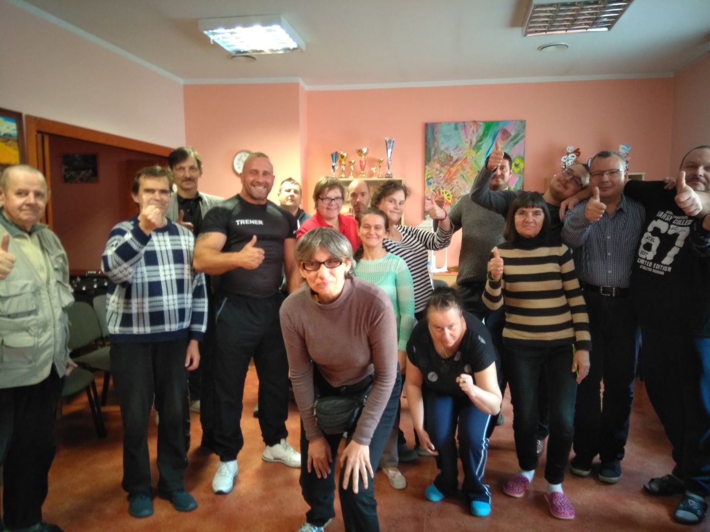 Mężczyzna sto i z grupą kobiet i mężczyzn, trzyma uniesioną dłoń z kciukiem do góry.