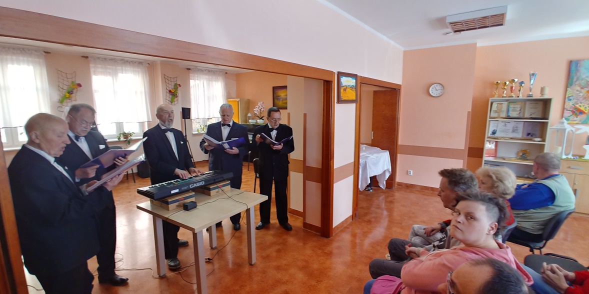 Starsi mężczyźni stoją przy stole ,ludzie siedzą na krzesełkach ,patrzą na nich.