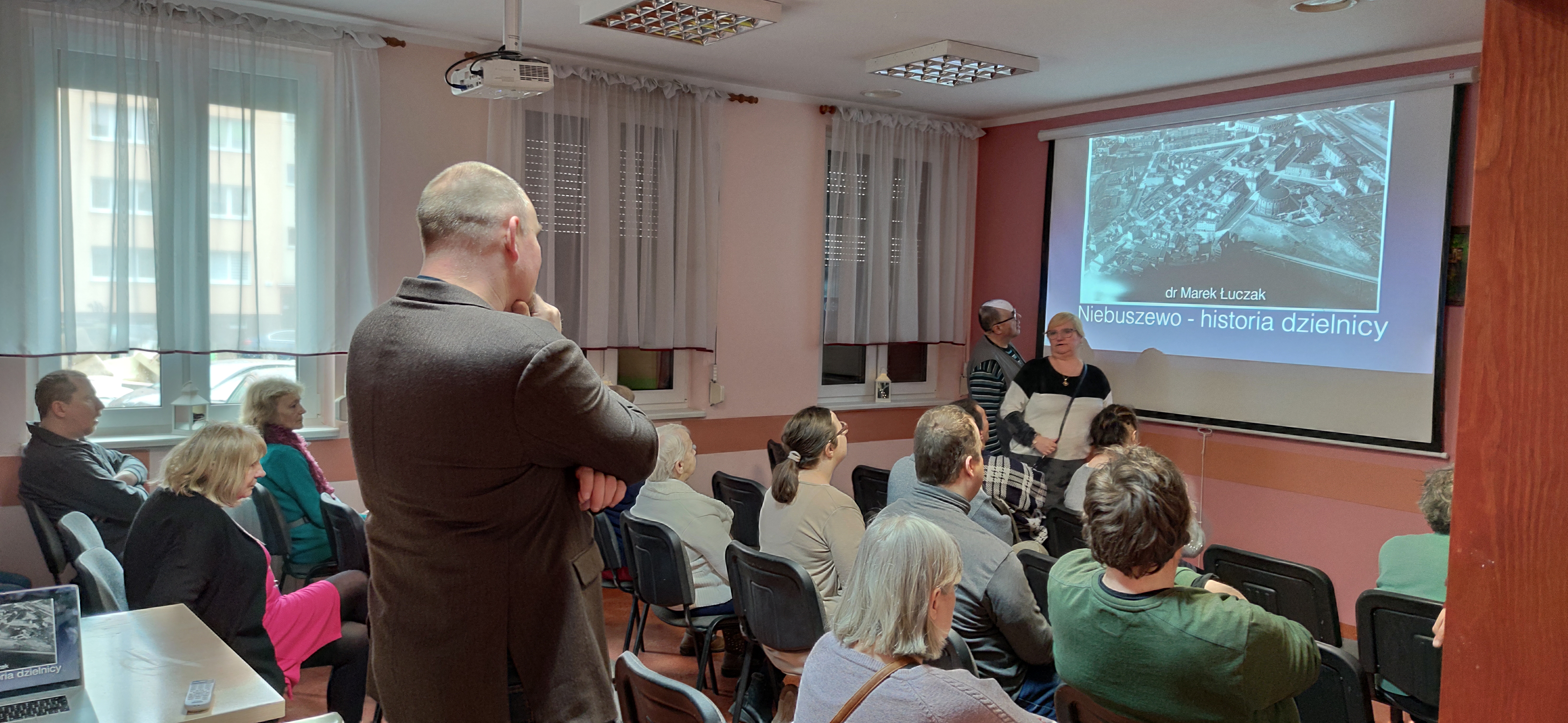 Grupa słuczaczy siedzi przed ekranem z tyłu prowadzący mówi.