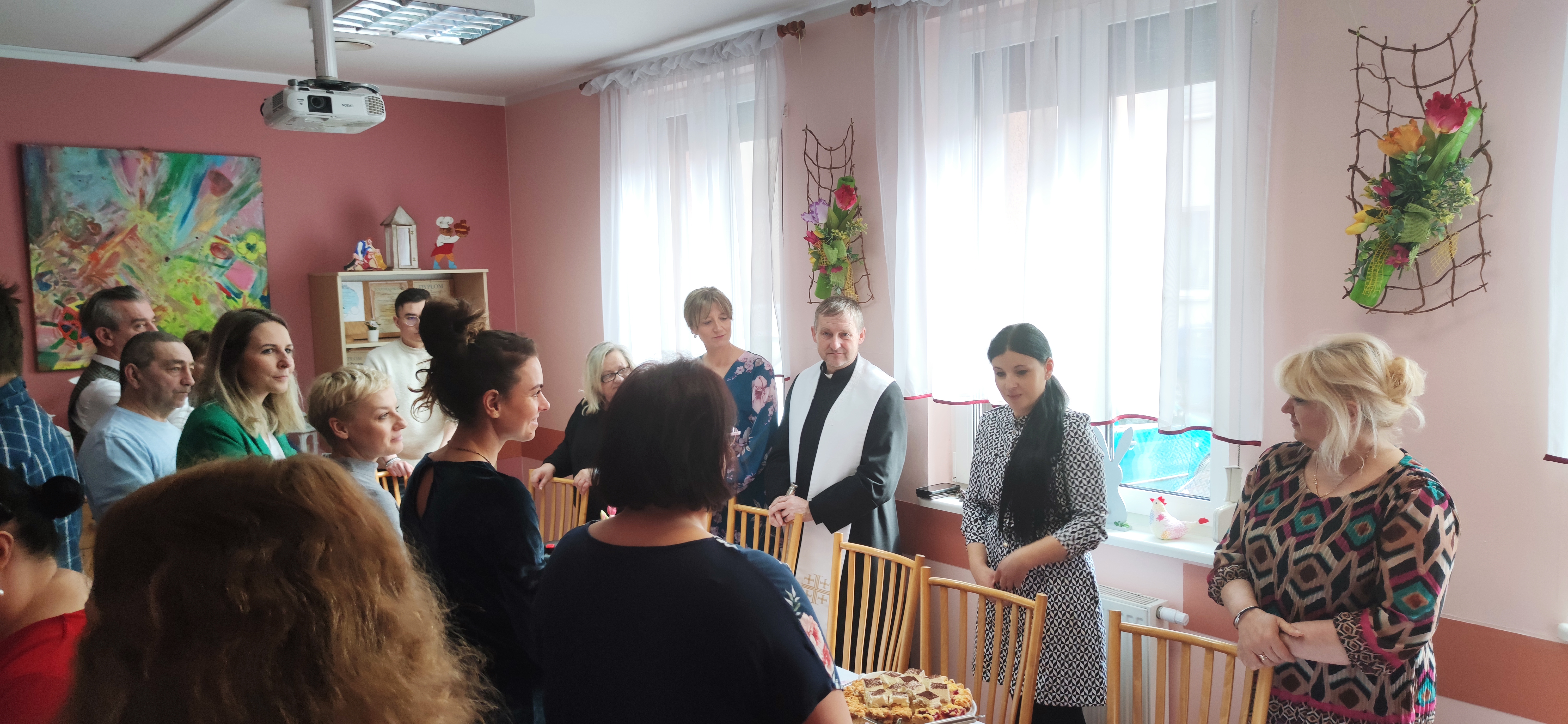 Grupa osób stoi z ksiądzem przy świątecznych stołach.