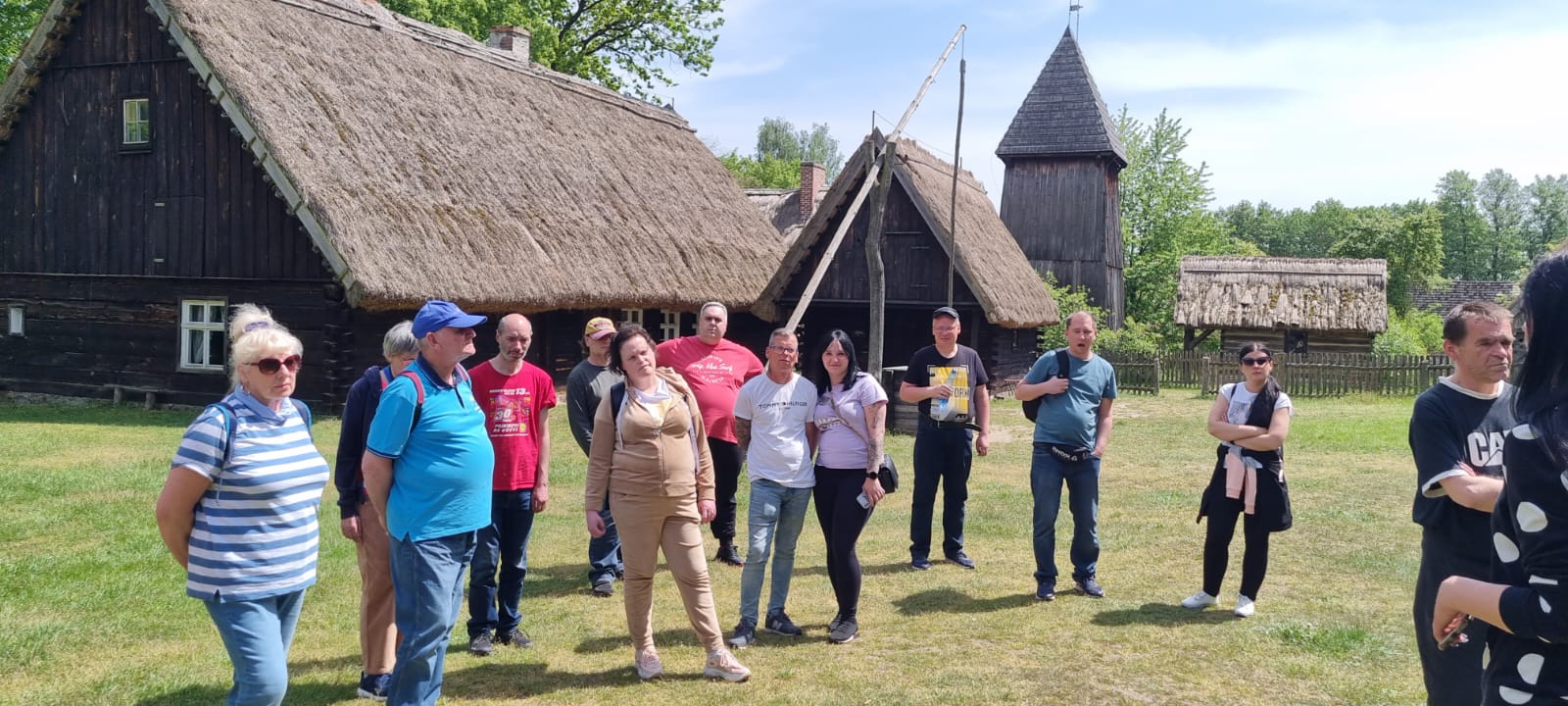 Grupa osób słucha przewodnika w wiejskim skansenie.
