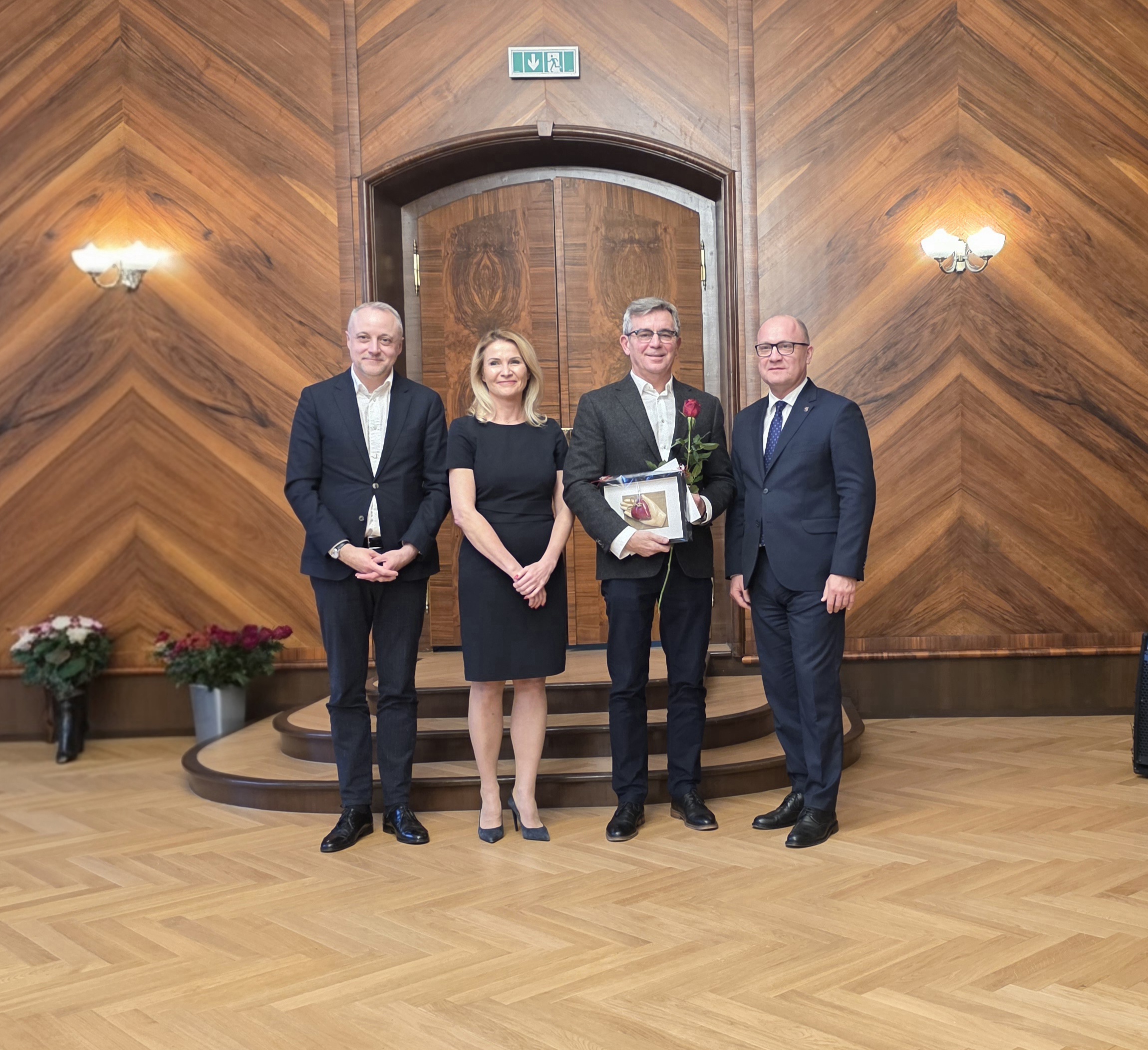 Cztery osoby , jedna z dyplomem i różą stoją w drewnianej sali