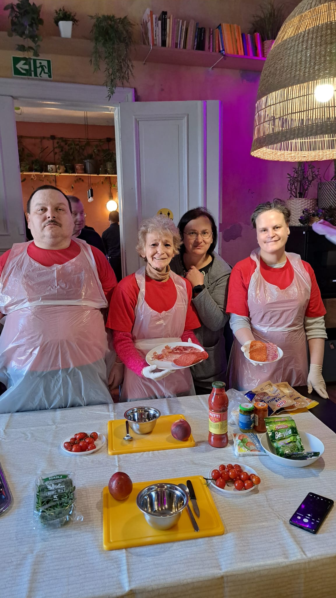 Grupa osób w koszulkach i fartuchach stoi nad stołem z przyborami kuchennymi.