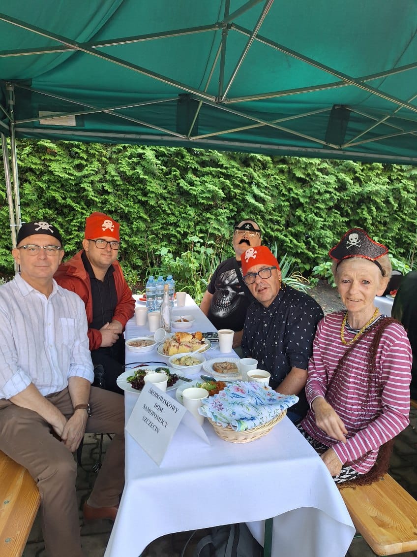 Osoby siedzą przy turystycznym stole pod namiotem.