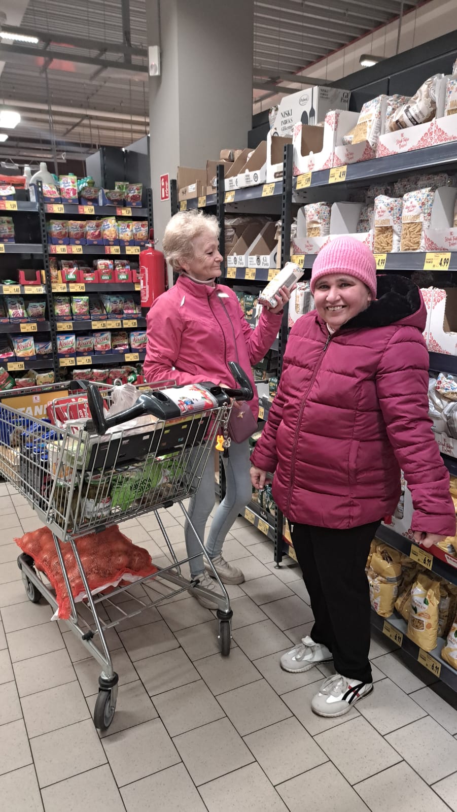 Dwie panie przy wózku sklepowym przeglądają towary na półkach.