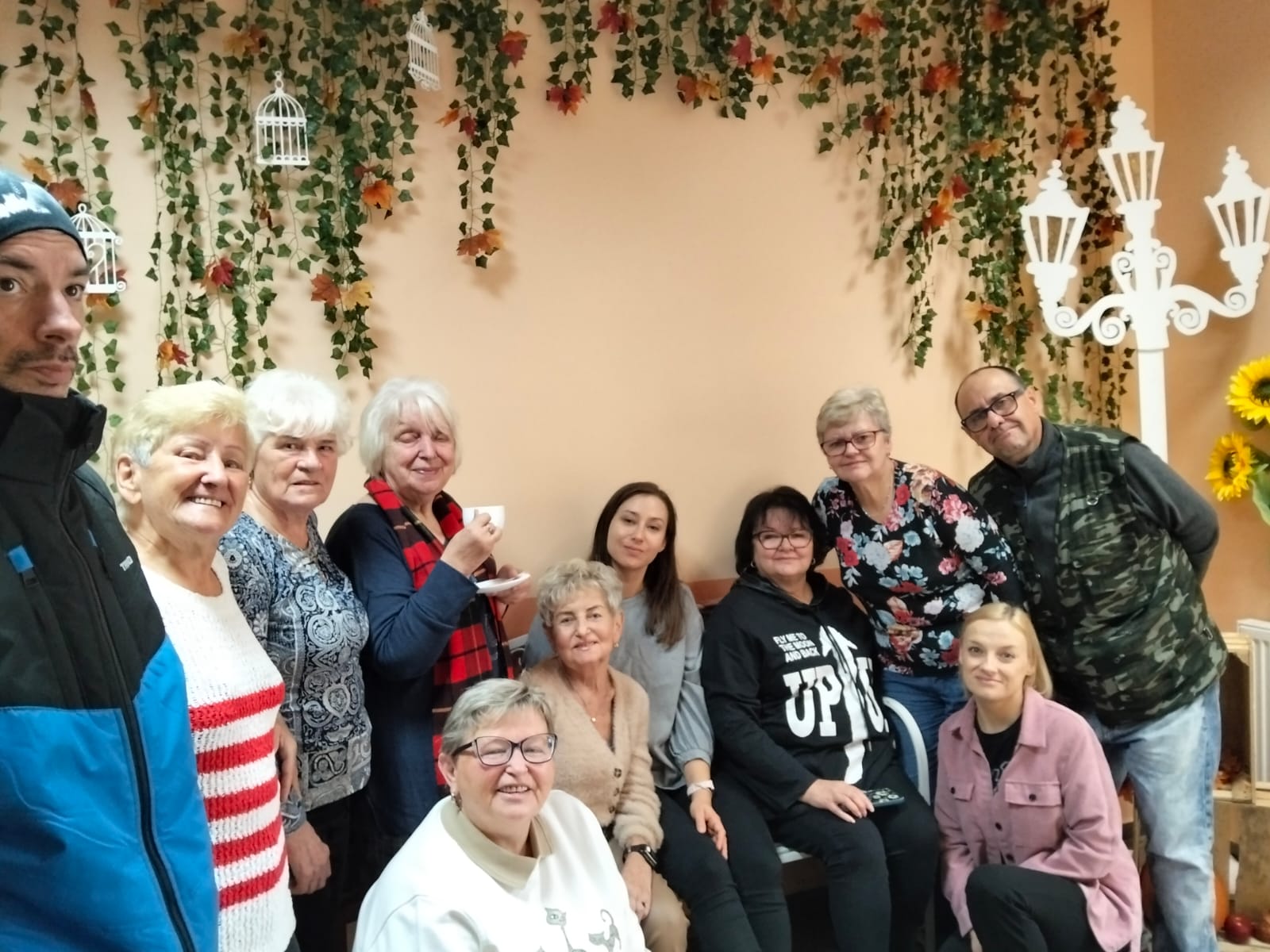 Grupa osób pozuje do zdjęcia pod jesienną dekoracją na ścianie.