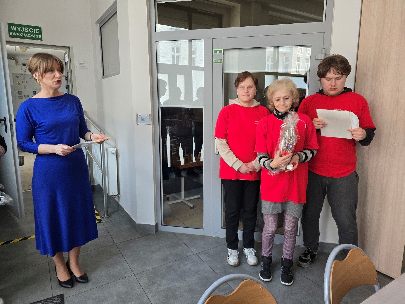 Kobieta w niebieskiej sukience, trzyma w dłoniach dyplom, który otrzymaj osoby w czerwonych koszulkach w tym dwie kobiety i jeden mężczyzna   .