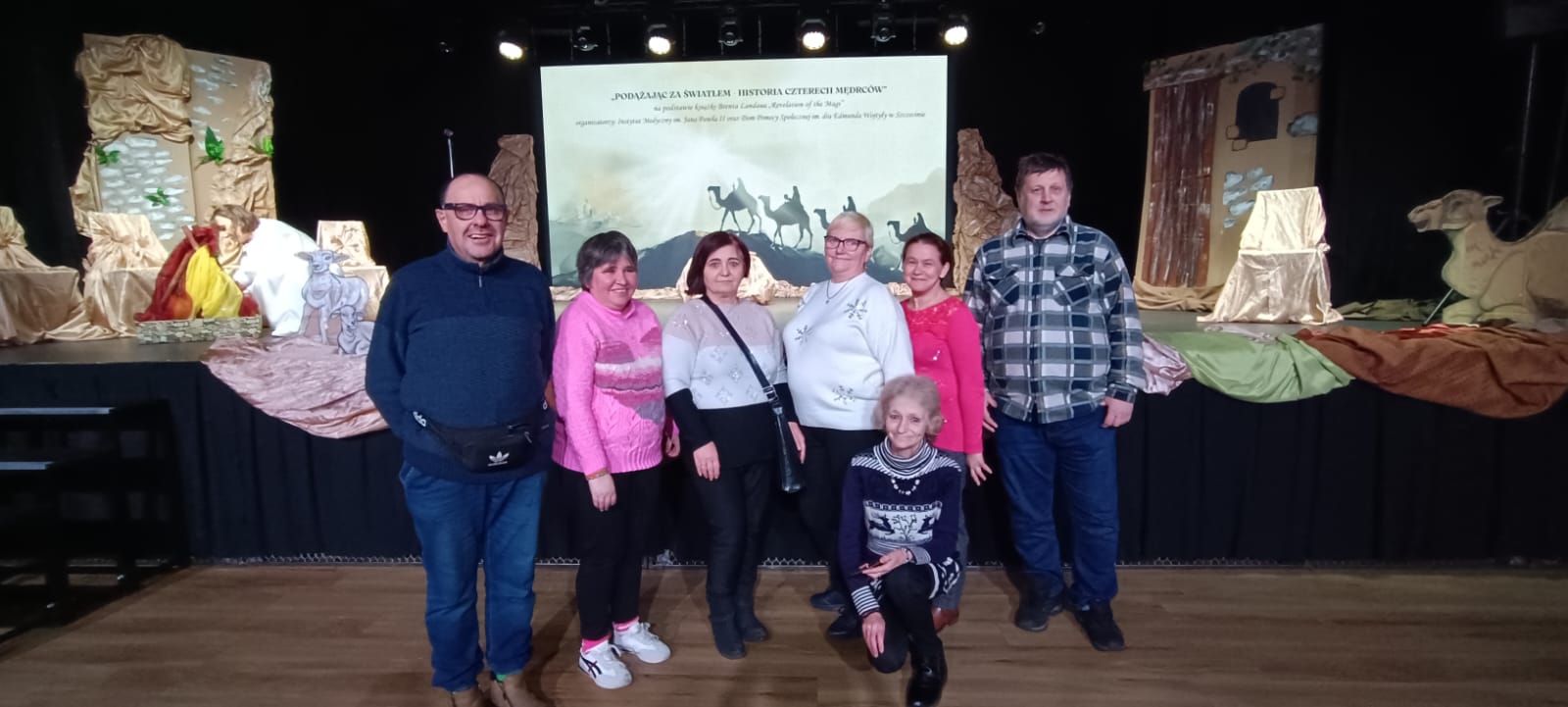 Grupa osób pozuje do zdjęcia przed sceną jasełek.