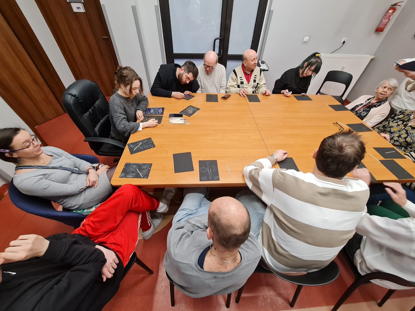 Grupa osób siedząca dookoła stołu na krzesłach. Na stole leżą czarne kartki.