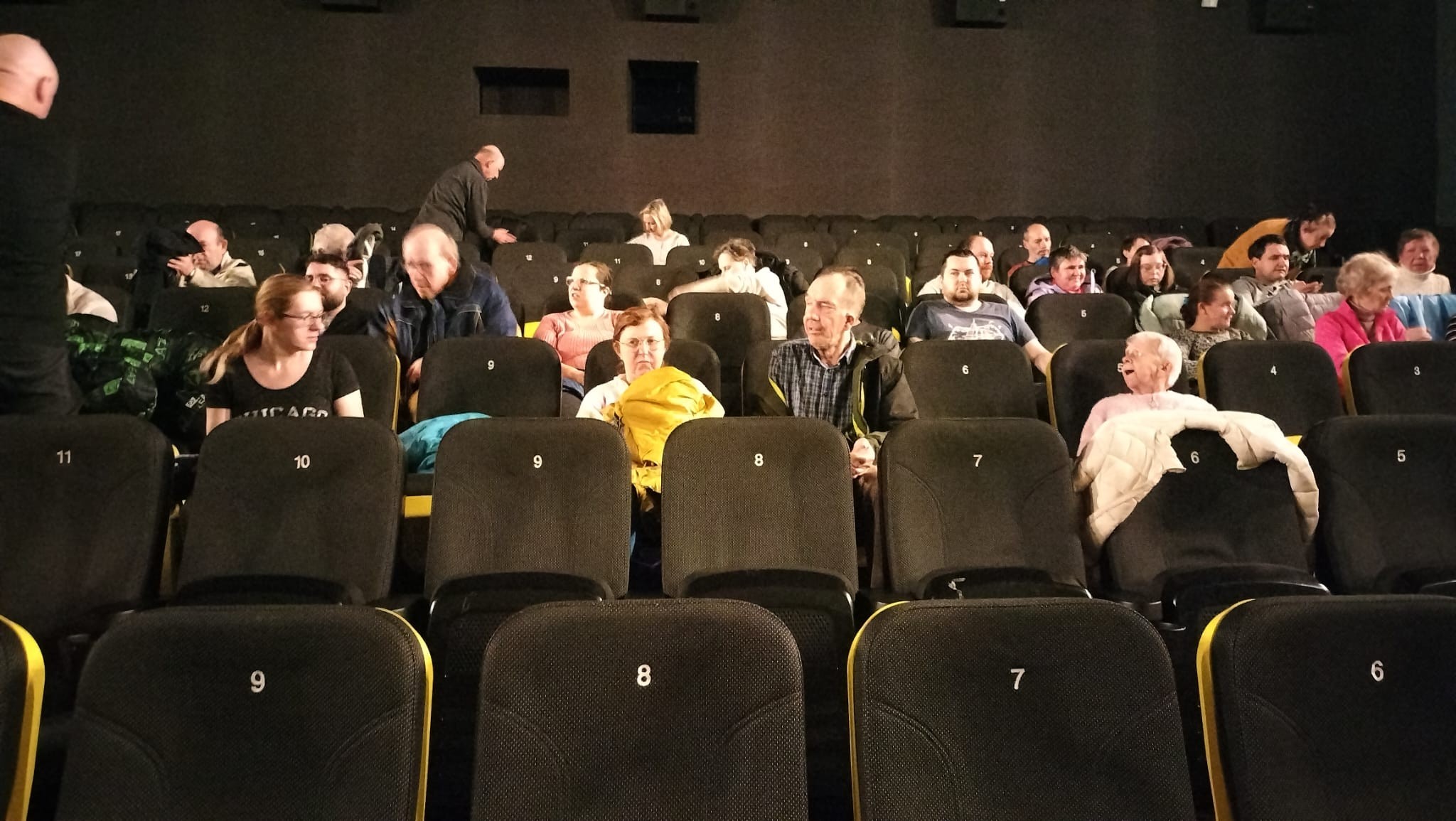 Grupa osób siedząca na czarno-żółtych fotelach w sali kinowej. 
