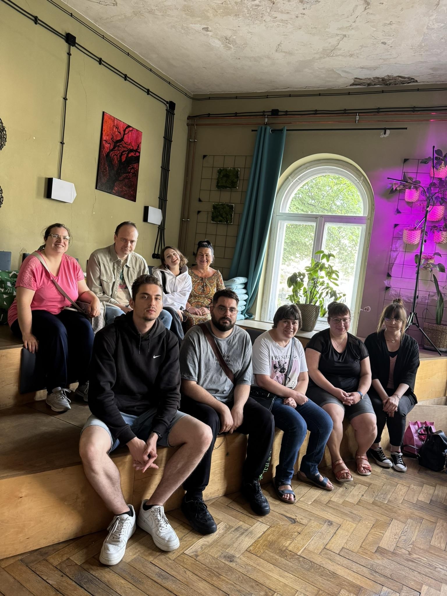 Grupa osób siedząca na schodach. W tle widać okno i zieloną roślinność.