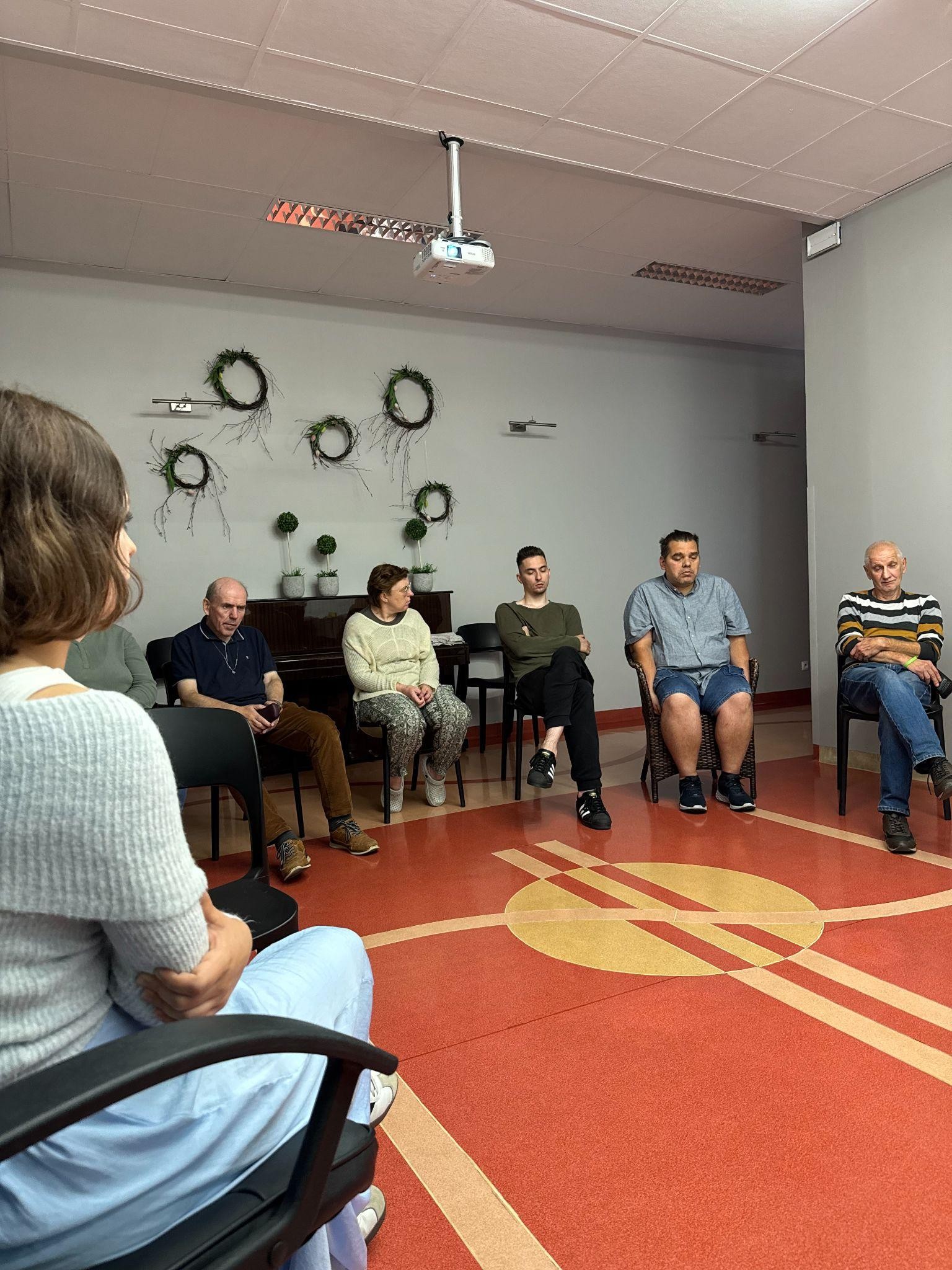 Grupa osób siedząca w półokręgu na krzesłach. W tle widać dekorację na ścianie i pianino.