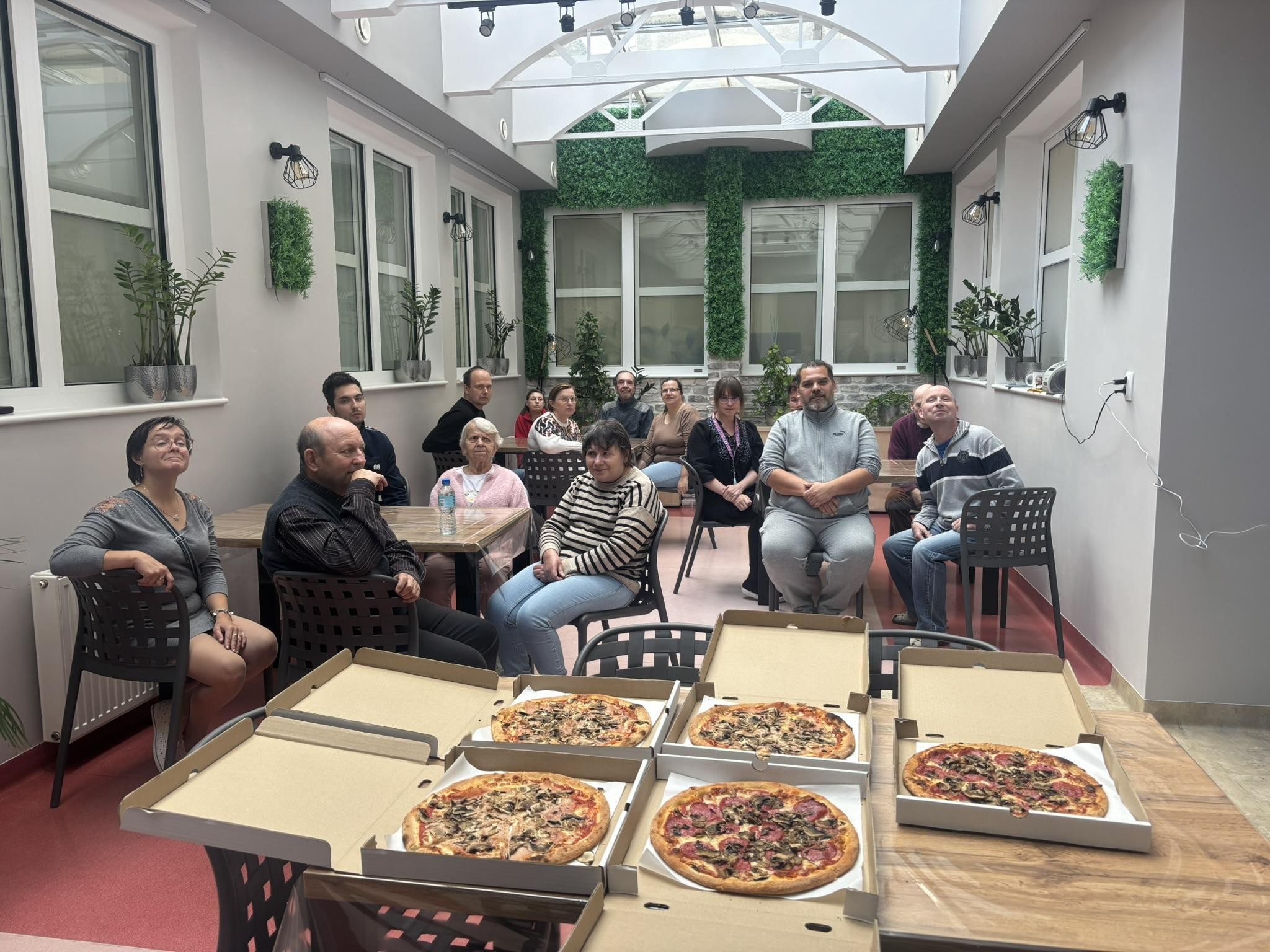 Grupa osób siedząca na krzesłach. Na pierwszym planie na stole widać 5 sztuk pizzy w kartonowych pojemnikach. W tle okna oraz zielone dekoracje.