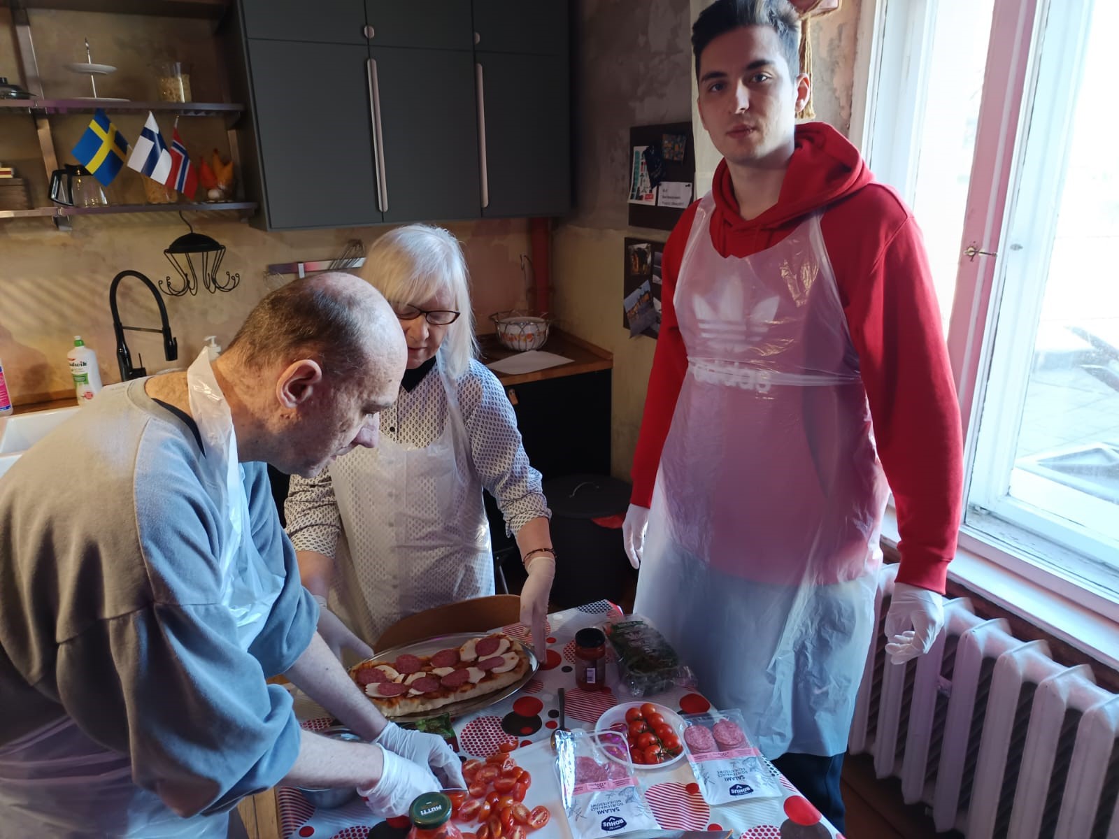 Trzy osoby w fartuchach komponujące potrawę. Na zdjęciu widoczny jest stół z produktami spożywczymi.