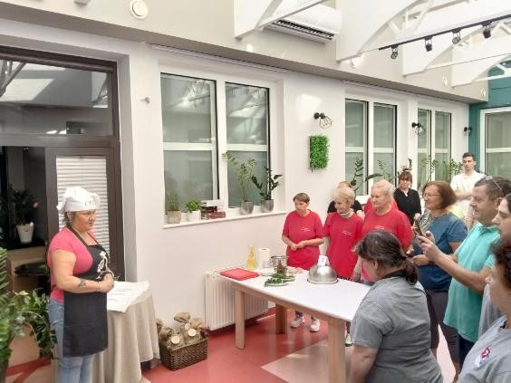 Grupa osób stojąca naprzeciwko osoby w czarnym fartuchu i białej czapce kucharskiej.