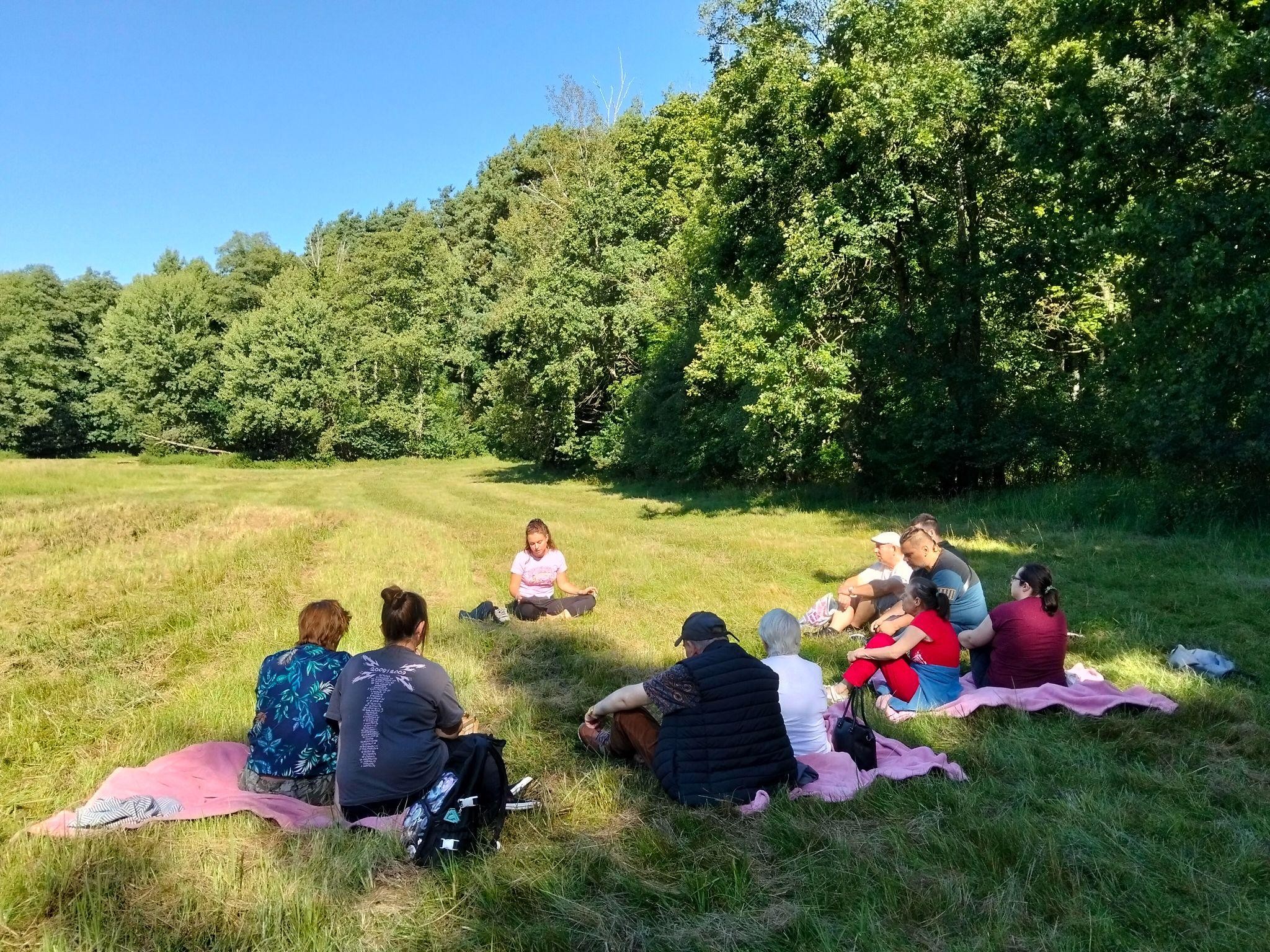 Grupa osób siedząca na trawie. Dookoła widać drzewa.