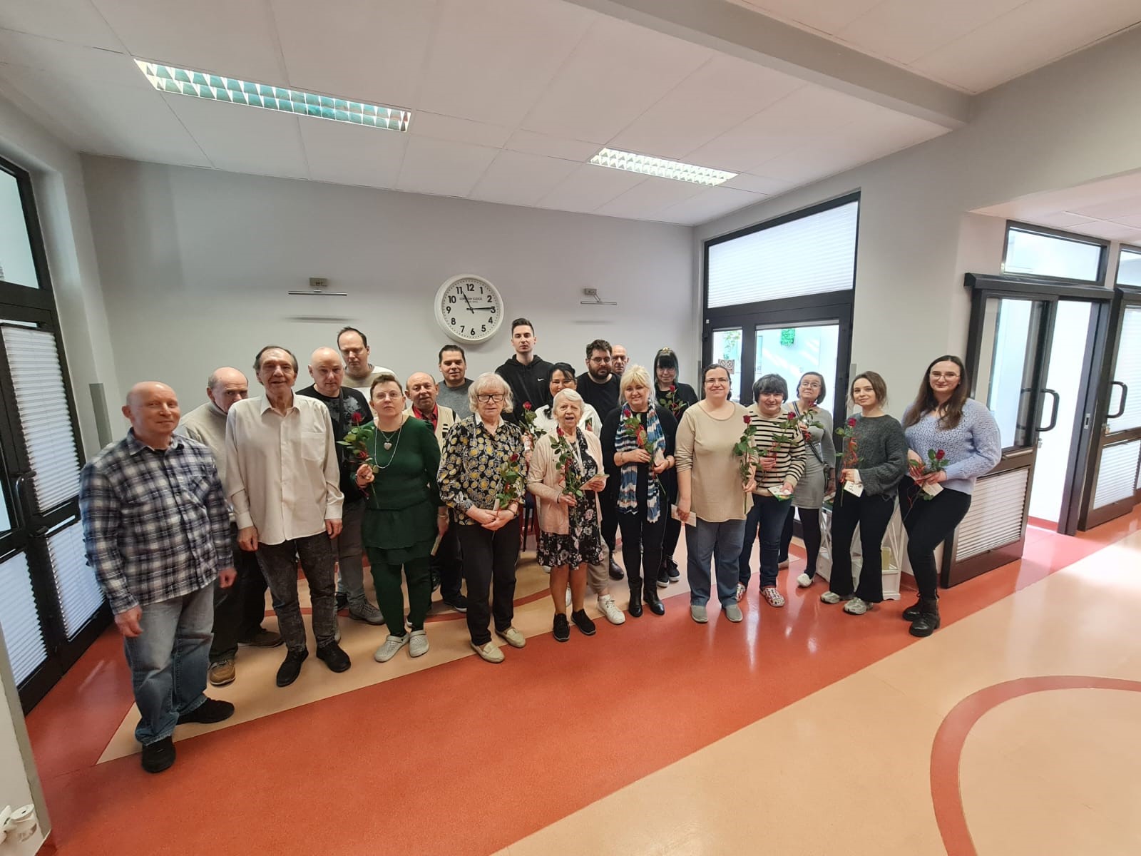 Grupa osób pozująca z różami. W tle widać drzwi oraz duży wiszący na ścianie zegar.
