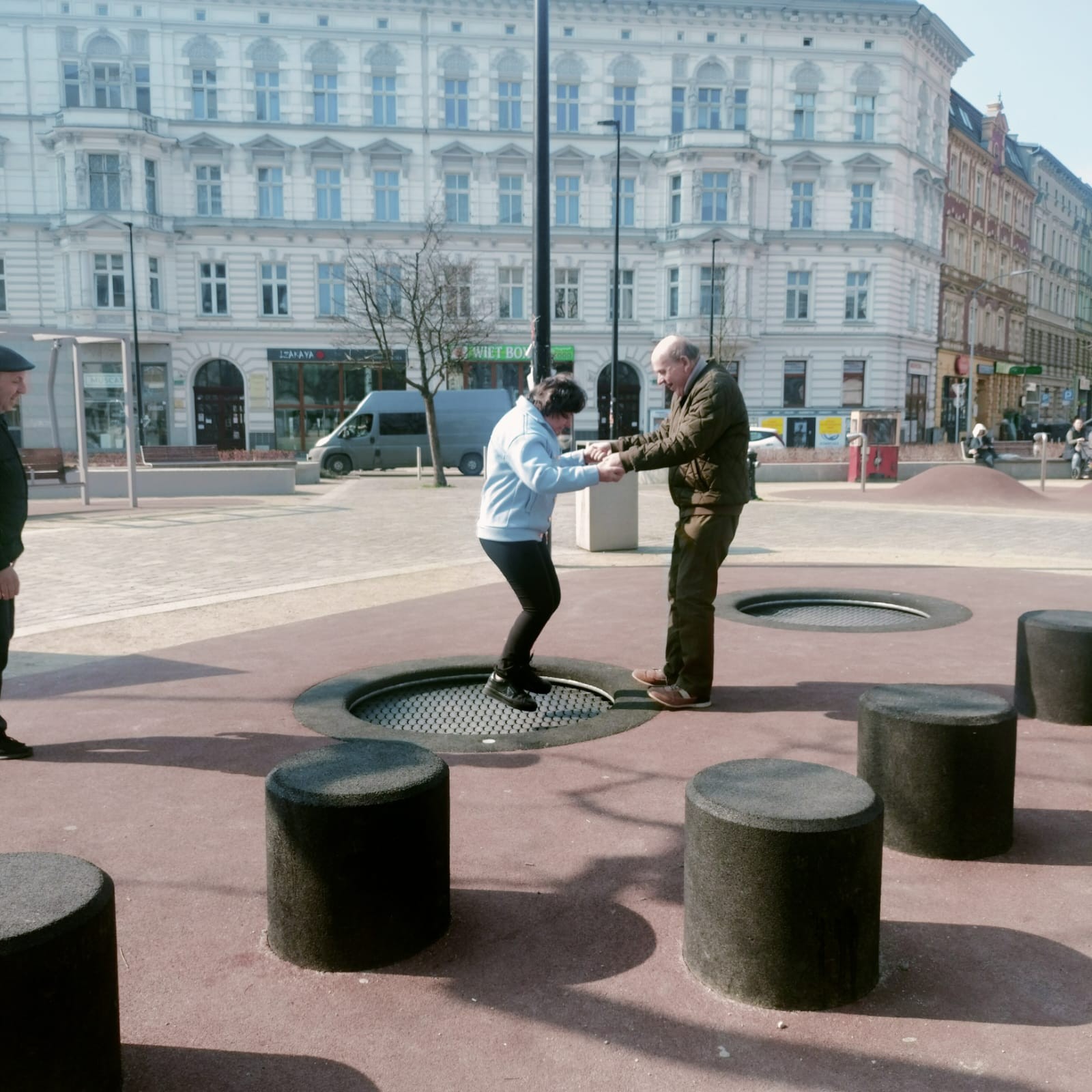 Dwie osoby trzymające się za ręce. Mężczyzna i kobieta korzystają z miejskich trampolin. W tle widać krajobraz miejski.