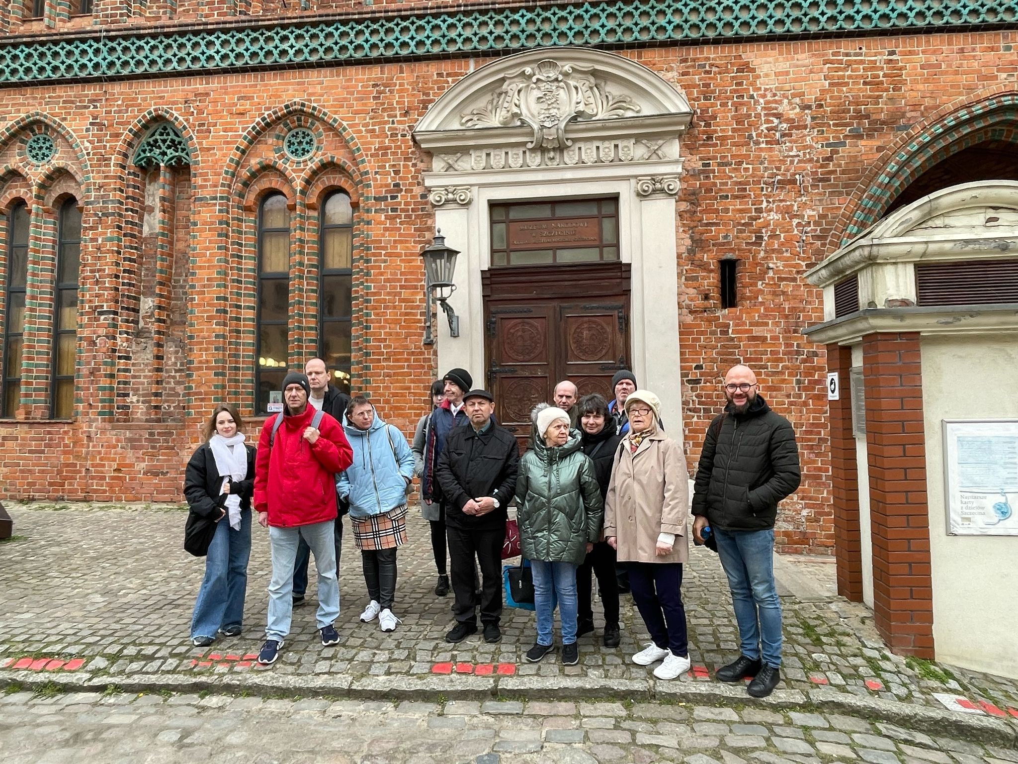 Grupa osób stojąca przed wejściem budynku z czerwonej cegły.