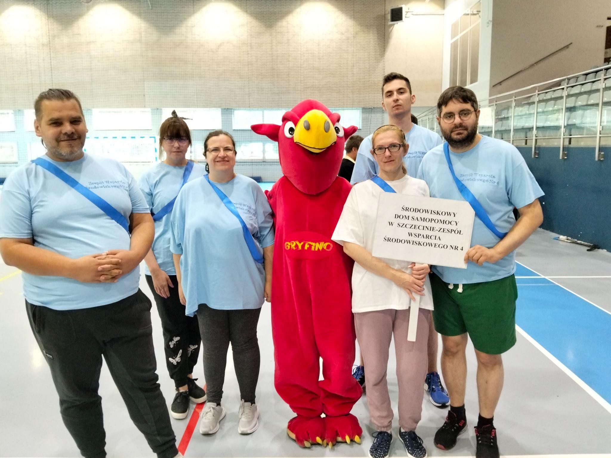 Grupa siedmiu osób pozująca do zdjęcia. Pięć osób ma błękitne koszulki, jedna ma na sobie białą a siódma jest w stroju czerwonego gryfa.