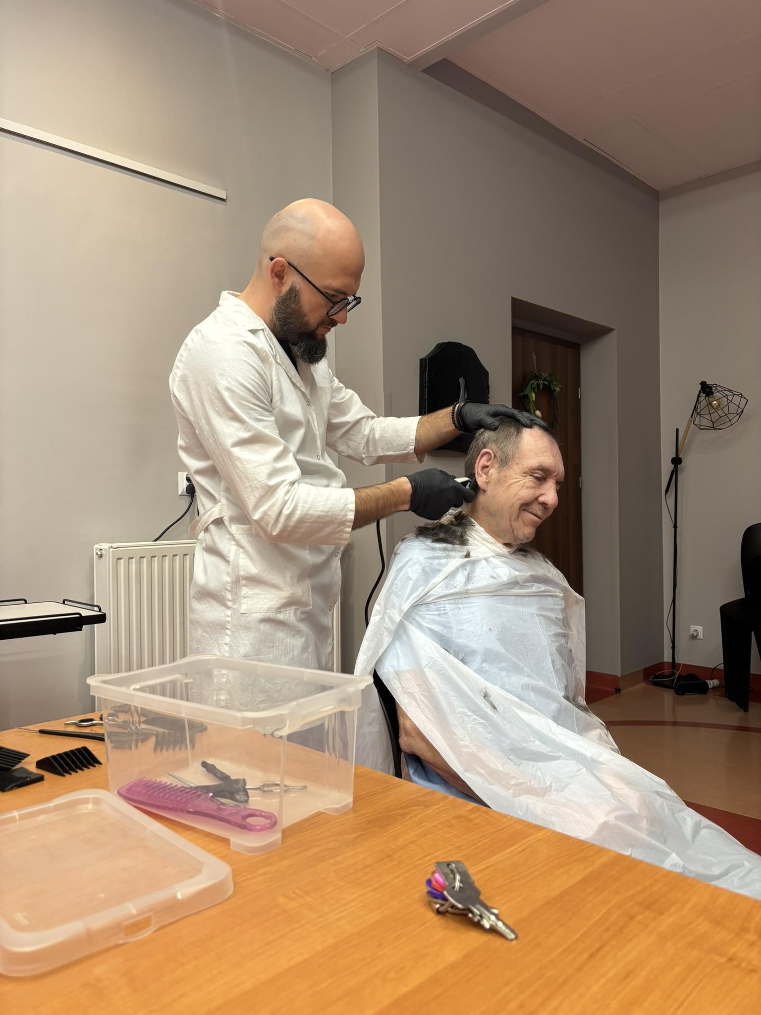 Na zdjęciu widać dwóch mężczyzn. Jeden w białym fartuchu ściana włosy drugiemu. Na stole widać pudełko z narzędziami fryzjerskimi.