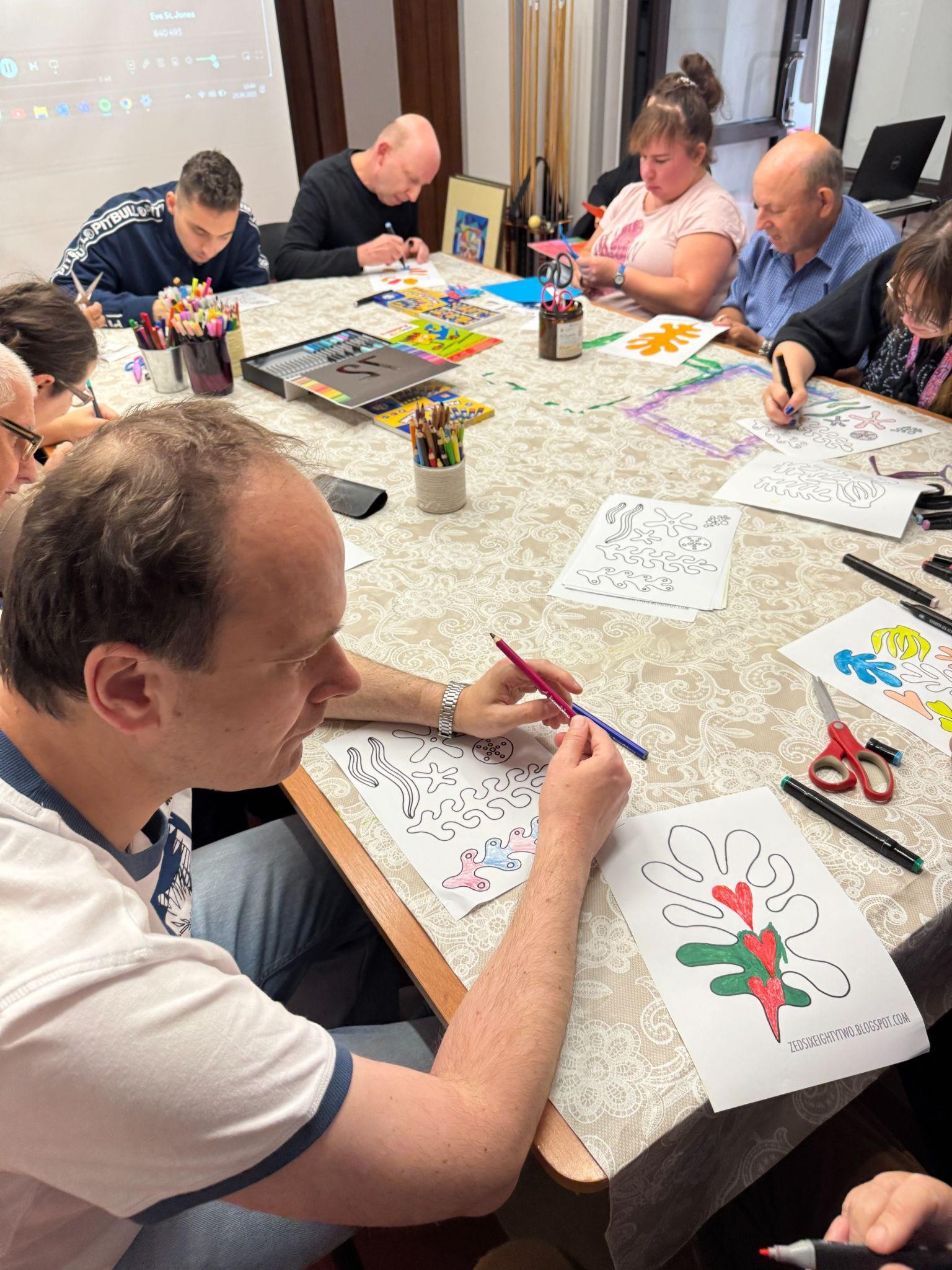 Grupa osób siedząca dookoła stołu. Osoby kolorują i rysują. Na stole widać kredki, mazaki, pastele oraz ceratę.
