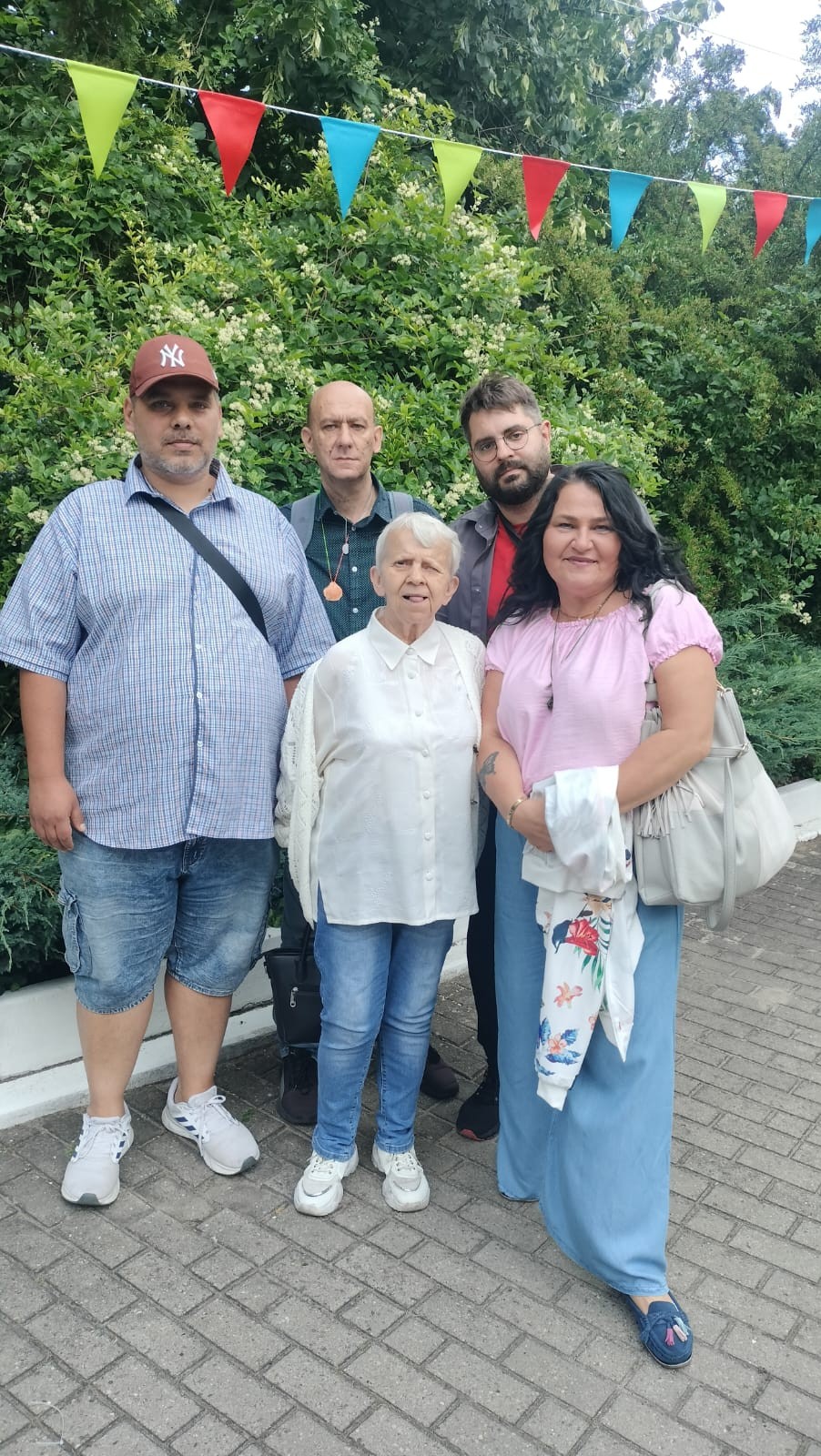Trzech mężczyzn i dwie kobiety stojące oraz pozujące do obiektywu aparatu. W tle widać kolorowe chorągiewki oraz zieleń.