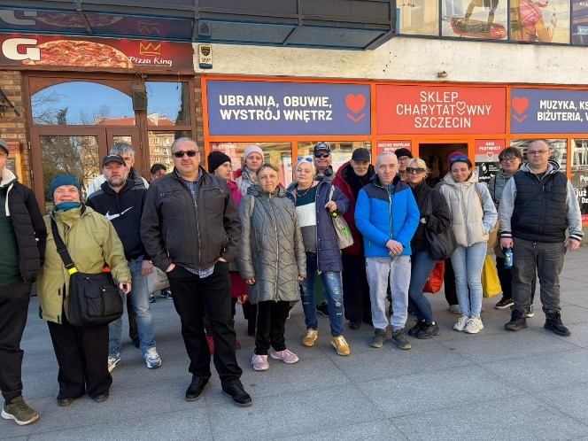 Grupa ok. 20 osób stoi przed sklepem i pozuje do zdjęcia. W tle napis „ SKLEP CHARYTATYWNY szczecin”.