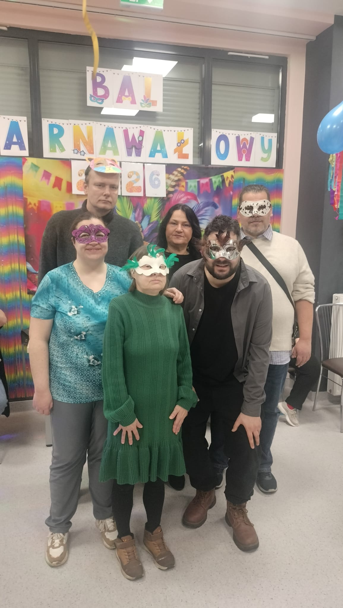 Grupa stojących osób. Cztery osoby mają na twarzach maski karnawałowe. W tle widać kolorowy napis i dekoracje.
